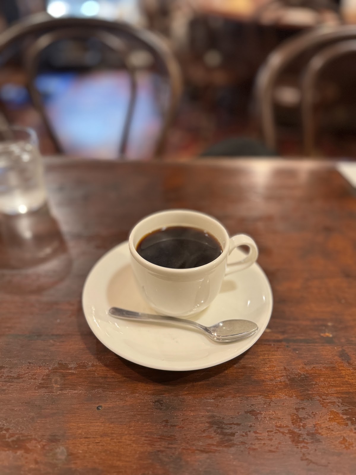 白い陶器カップに注がれたブラックコーヒーと銀のスプーン、背後にぼけた木製チェア