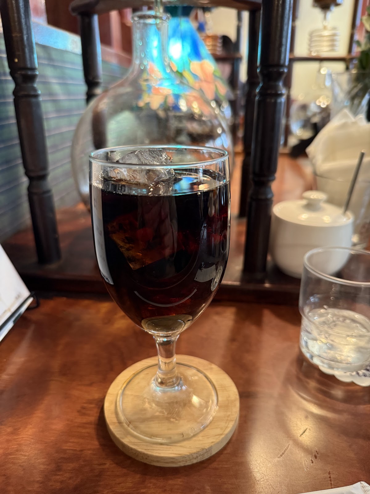 ワイングラスに注がれた黒いアイスコーヒーと氷、背後にステンドグラス風の青いランプと水出し器が並ぶ店内